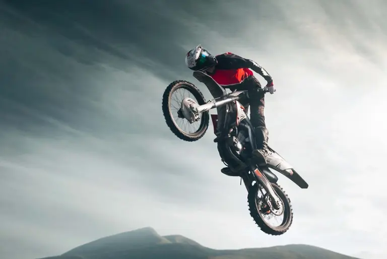 A motocross rider in red and black gear performs a high jump against a cloudy sky. The image conveys action and thrill, with distant hills below.